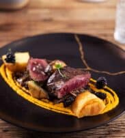 A gourmet dish featuring sliced medium-rare steak on a bed of orange purée, garnished with blackberries, microgreens, mushrooms, and toasted bread on a black plate.