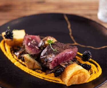 A gourmet dish featuring sliced medium-rare steak on a bed of orange purée, garnished with blackberries, microgreens, mushrooms, and toasted bread on a black plate.