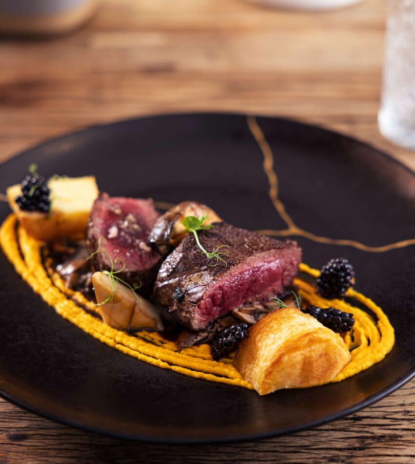 A gourmet dish featuring sliced medium-rare steak on a bed of orange purée, garnished with blackberries, microgreens, mushrooms, and toasted bread on a black plate.