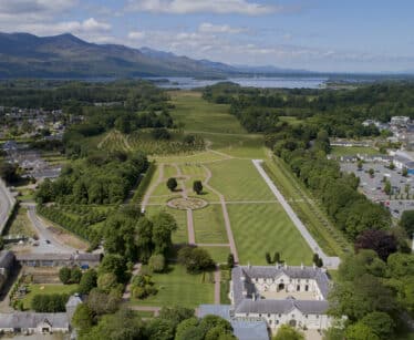 Aerial view of a formal garden with symmetrical paths, surrounded by trees, buildings, and a town; mountains and a lake are visible in the background under a partly cloudy sky.