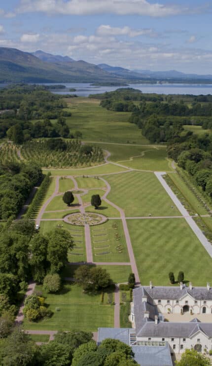 Aerial view of a formal garden with symmetrical paths, surrounded by trees, buildings, and a town; mountains and a lake are visible in the background under a partly cloudy sky.