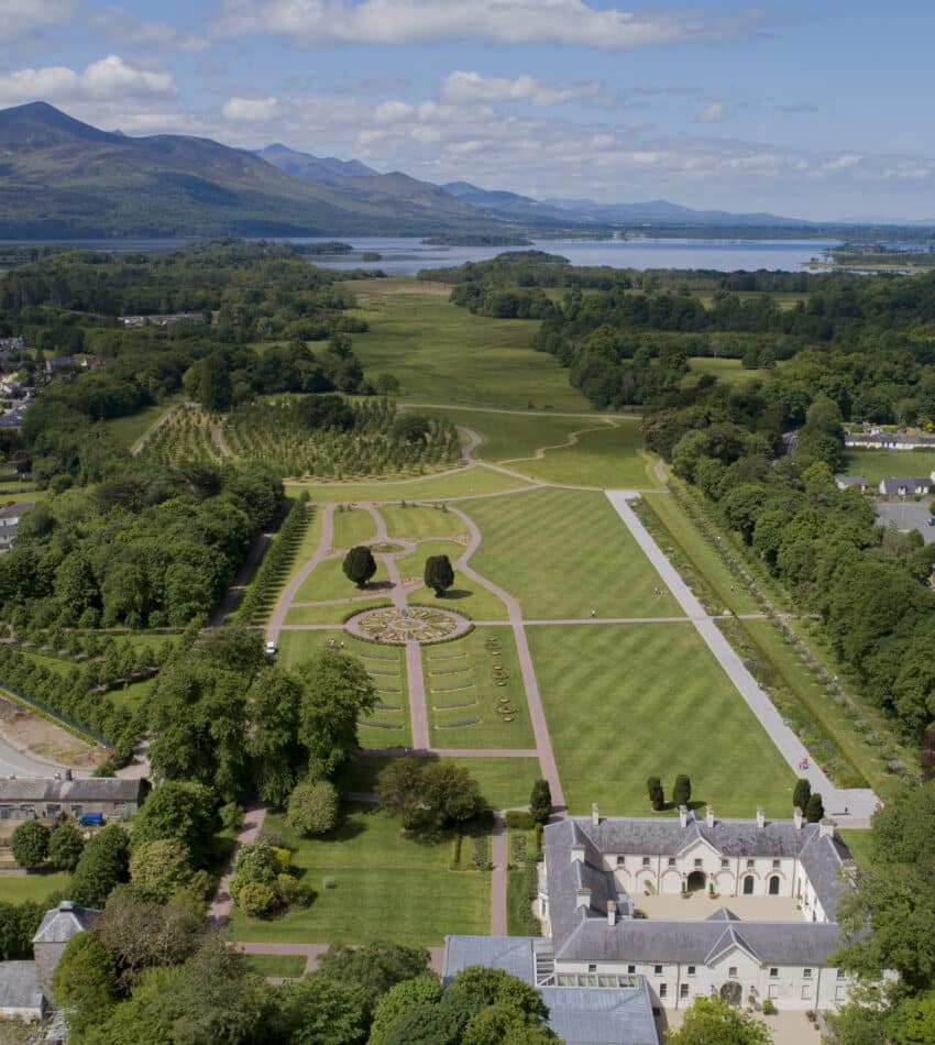 Aerial view of a formal garden with symmetrical paths, surrounded by trees, buildings, and a town; mountains and a lake are visible in the background under a partly cloudy sky.