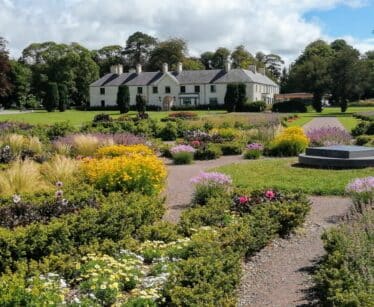 A large house with a grey roof sits in the background, surrounded by well-kept gardens with colourful flowers and shrubs under a partly cloudy sky.