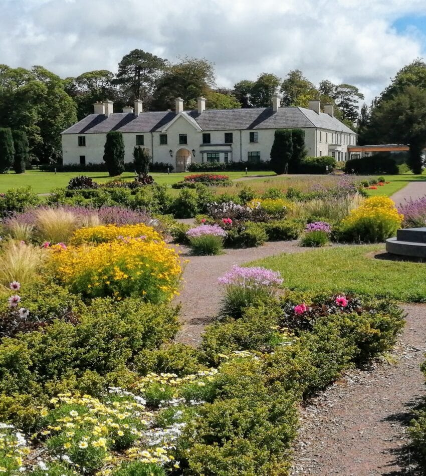A large house with a grey roof sits in the background, surrounded by well-kept gardens with colourful flowers and shrubs under a partly cloudy sky.