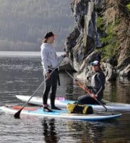 Two people on paddleboards, one standing and one kneeling, talk near a rocky shore with trees and hills in the background.