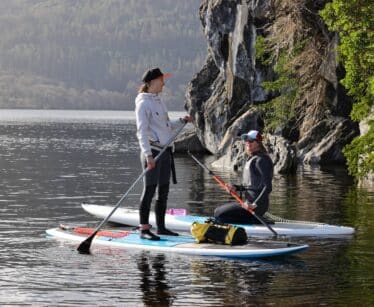 Two people on paddleboards, one standing and one kneeling, talk near a rocky shore with trees and hills in the background.