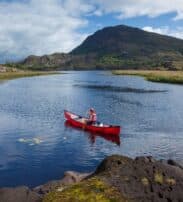 A person in a red canoe paddles on a calm river surrounded by grassy banks and hills under a partly cloudy sky.
