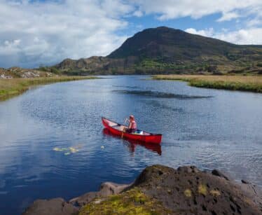 A person in a red canoe paddles on a calm river surrounded by grassy banks and hills under a partly cloudy sky.