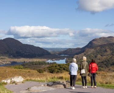 Three people in jackets stand on a path overlooking a scenic valley with mountains, lakes, and a partly cloudy sky in the background.