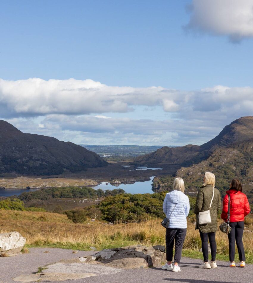 Three people in jackets stand on a path overlooking a scenic valley with mountains, lakes, and a partly cloudy sky in the background.