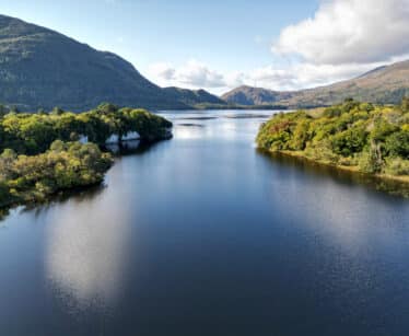 A calm lake stretches between two forested hills under a partly cloudy sky, with distant mountains visible in the background.