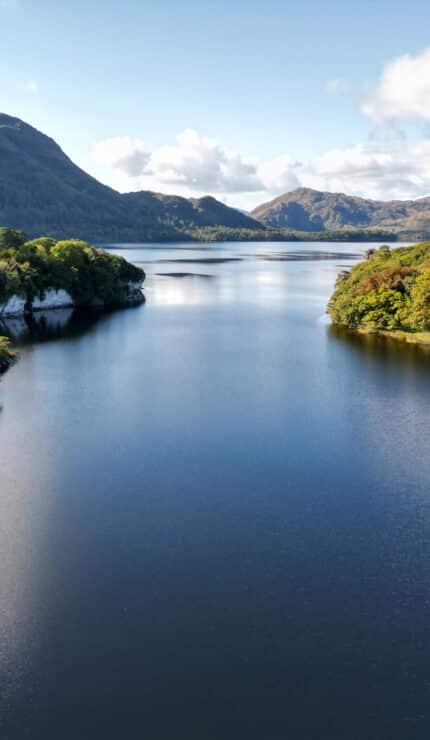 A calm lake stretches between two forested hills under a partly cloudy sky, with distant mountains visible in the background.