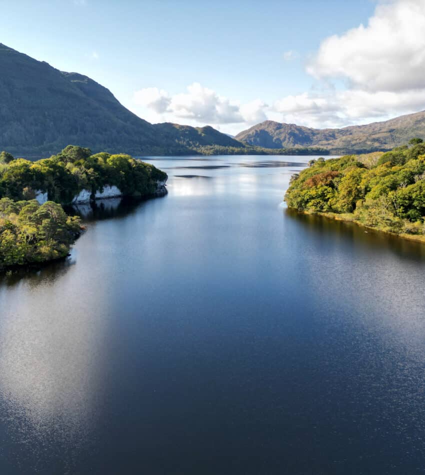 A calm lake stretches between two forested hills under a partly cloudy sky, with distant mountains visible in the background.