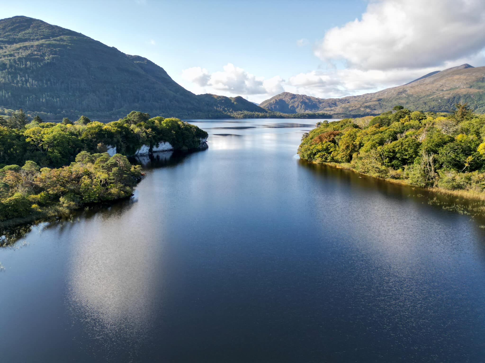 A calm lake stretches between two forested hills under a partly cloudy sky, with distant mountains visible in the background.