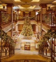 Grand hotel lobby decorated for Christmas with a large, ornate tree at the centre, garlands with lights on stair railings, and seating areas on both sides.