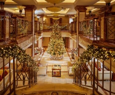 Grand hotel lobby decorated for Christmas with a large, ornate tree at the centre, garlands with lights on stair railings, and seating areas on both sides.