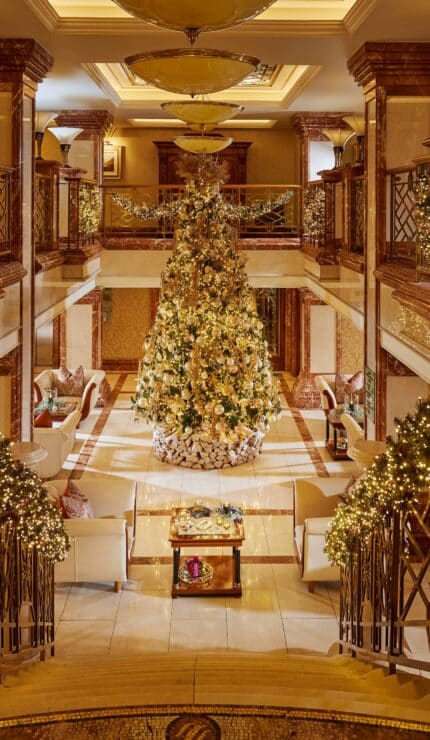 Grand hotel lobby decorated for Christmas with a large, ornate tree at the centre, garlands with lights on stair railings, and seating areas on both sides.