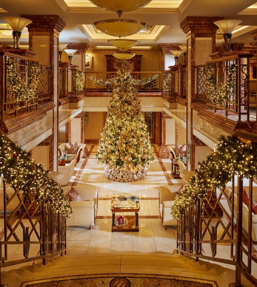 Grand hotel lobby decorated for Christmas with a large, ornate tree at the centre, garlands with lights on stair railings, and seating areas on both sides.