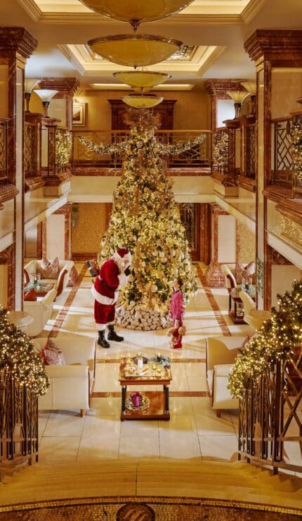 A large, decorated Christmas tree stands in the centre of a lavishly decorated hotel lobby with Father Christmas and a child nearby. Holiday lights and garlands adorn the staircase and railings.