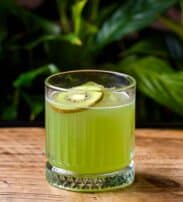 A glass of green kiwi juice garnished with a slice of fresh kiwi, placed on a wooden surface with green plants in the background.
