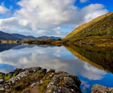 A person in a blue jacket stands on rocks by a calm lake with mountains and clouds reflected in the water under a partly cloudy sky.