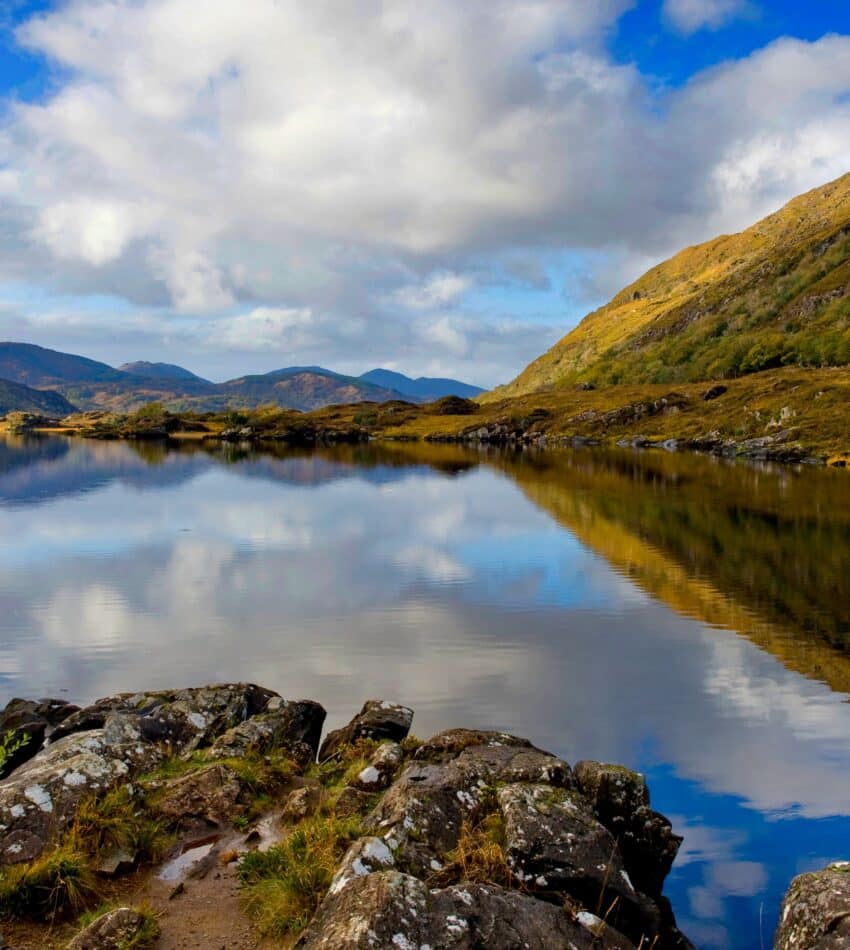 A person in a blue jacket stands on rocks by a calm lake with mountains and clouds reflected in the water under a partly cloudy sky.