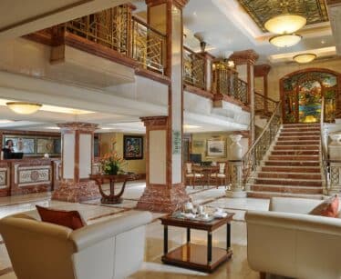 Elegant hotel lobby with marble floors, beige sofas, a coffee table, large staircase, reception desk, and decorative lighting. Ornate railings and stained glass accent the upper level.