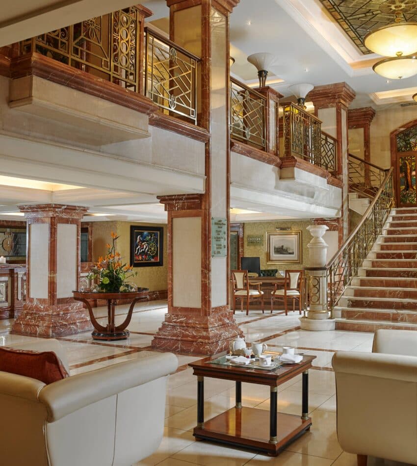 Elegant hotel lobby with marble floors, beige sofas, a coffee table, large staircase, reception desk, and decorative lighting. Ornate railings and stained glass accent the upper level.