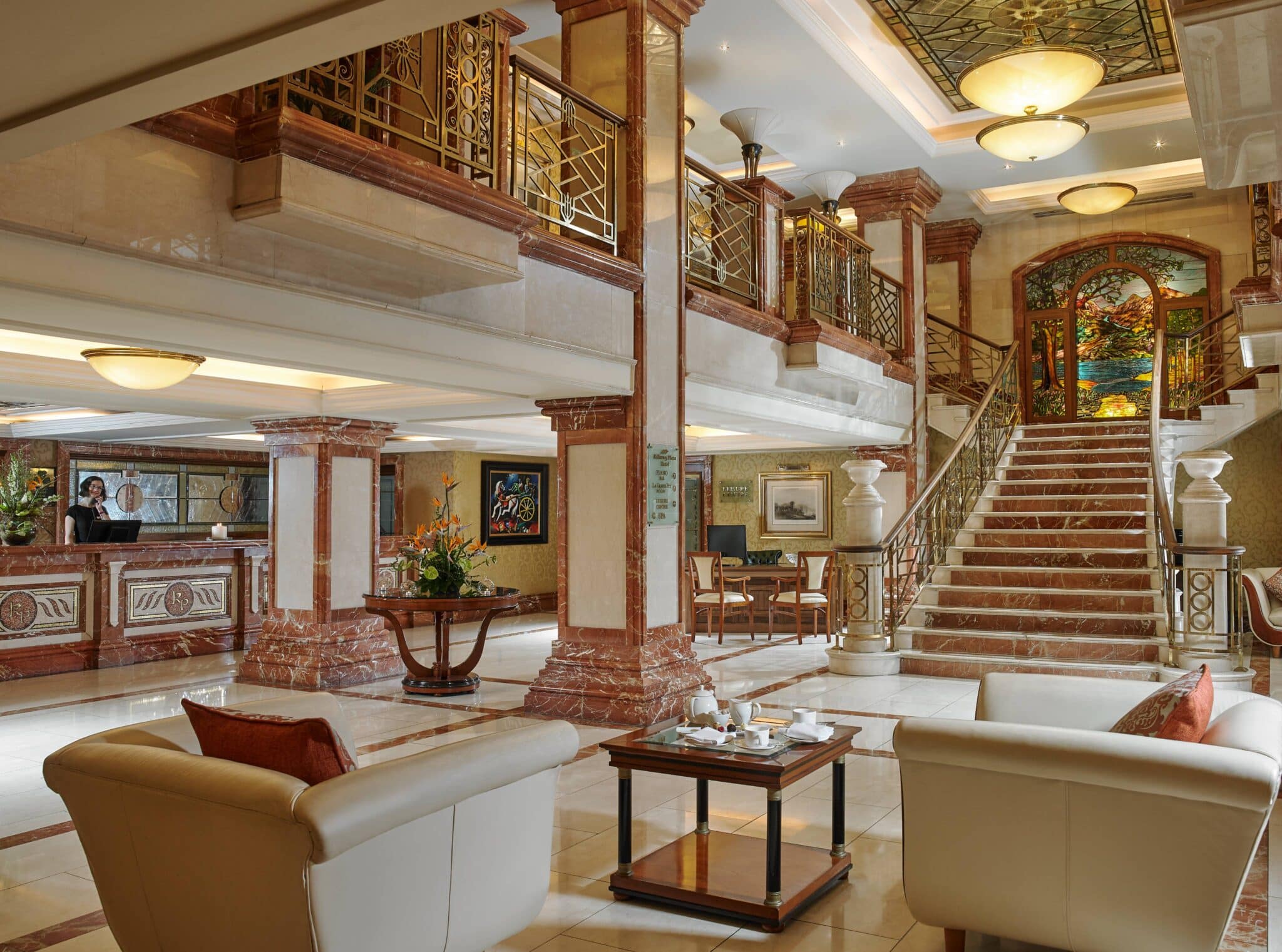Elegant hotel lobby with marble floors, beige sofas, a coffee table, large staircase, reception desk, and decorative lighting. Ornate railings and stained glass accent the upper level.