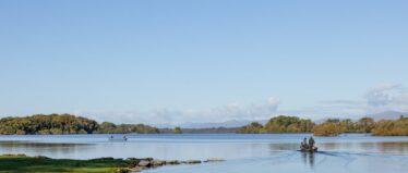 A small boat with people moves across a calm lake under a clear blue sky, with trees and distant hills in the background.