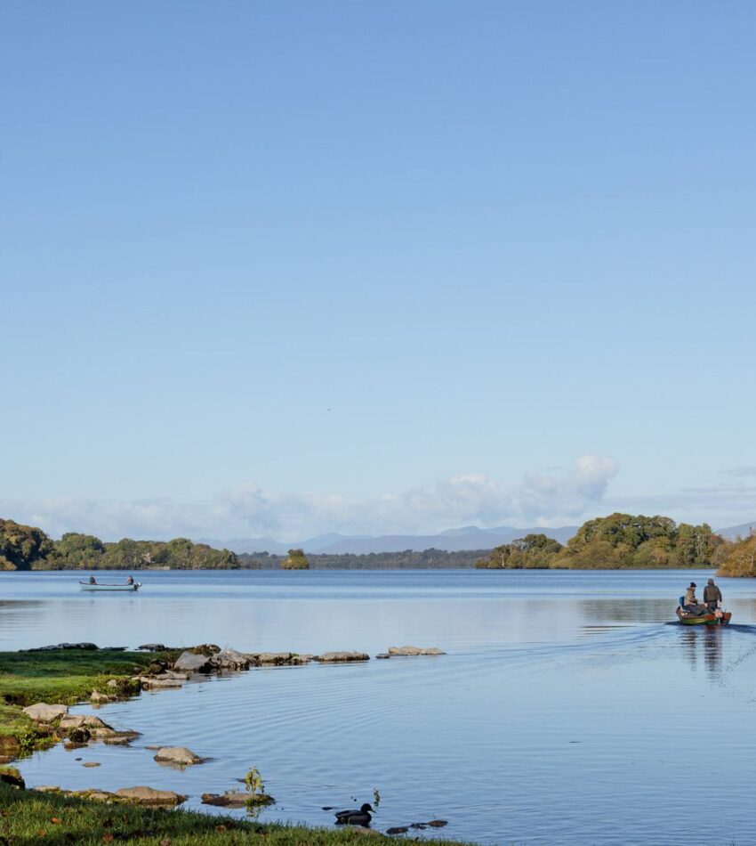 A small boat with people moves across a calm lake under a clear blue sky, with trees and distant hills in the background.