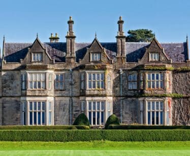A large, historic stone mansion with tall chimneys, many windows, ivy-covered sections, and neatly trimmed hedges in front, set against a clear blue sky.
