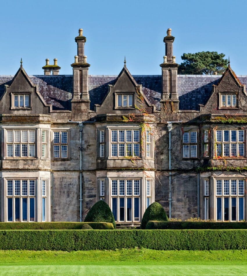 A large, historic stone mansion with tall chimneys, many windows, ivy-covered sections, and neatly trimmed hedges in front, set against a clear blue sky.