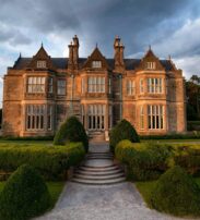 A large, historic stone manor with multiple chimneys and tall windows, surrounded by manicured hedges and gardens under a partly cloudy sky.