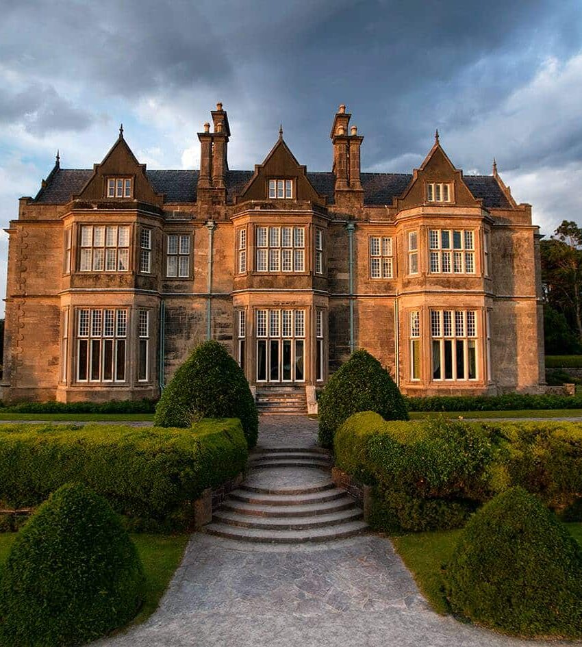 A large, historic stone manor with multiple chimneys and tall windows, surrounded by manicured hedges and gardens under a partly cloudy sky.