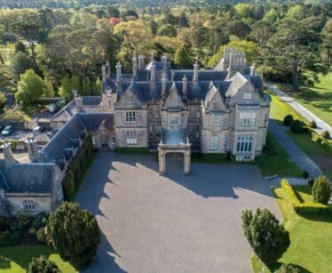 Aerial view of a large, historic stone mansion with multiple chimneys and gabled roofs, surrounded by manicured lawns and trees.