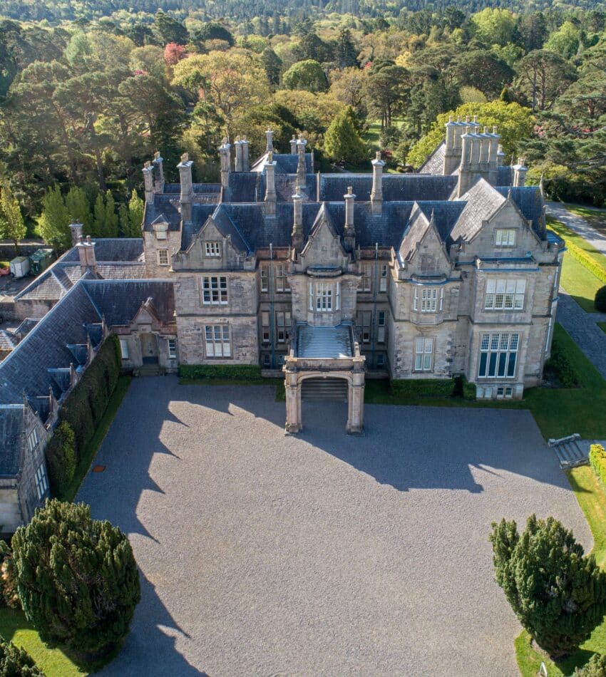 Aerial view of a large, historic stone mansion with multiple chimneys and gabled roofs, surrounded by manicured lawns and trees.