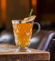 A clear glass mug filled with an amber-coloured drink, garnished with a sprig of rosemary and a dried apple slice, sits on a wooden table.