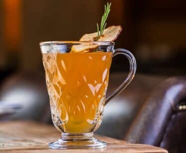 A clear glass mug filled with an amber-coloured drink, garnished with a sprig of rosemary and a dried apple slice, sits on a wooden table.