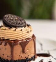 A chocolate mousse dessert with chocolate drizzle, biscuit crumb base, and a chocolate sandwich biscuit on top, served on a white plate.