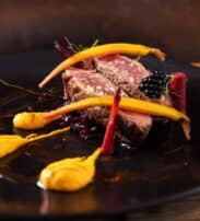 A gourmet dish featuring slices of seared beef, baby carrots, yellow purée, red vegetable garnish, and a blackberry on a black plate.