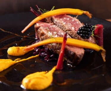 A gourmet dish featuring slices of seared beef, baby carrots, yellow purée, red vegetable garnish, and a blackberry on a black plate.