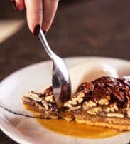 A hand holding a spoon scooping a slice of pecan pie with a scoop of ice cream on a white plate.