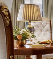A round wooden table with a lamp, a vase of flowers, a book, and a plate of pastries, next to tufted chairs and a window with curtains.