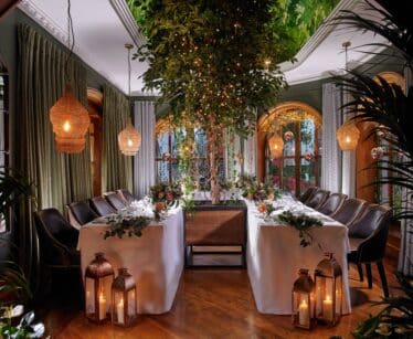 Elegant dining room with two tables arranged in an L-shape, decorated with greenery, candles, and lanterns, featuring a large indoor tree and ambient hanging lights.