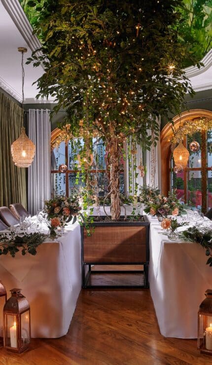 Elegant dining room with two tables arranged in an L-shape, decorated with greenery, candles, and lanterns, featuring a large indoor tree and ambient hanging lights.