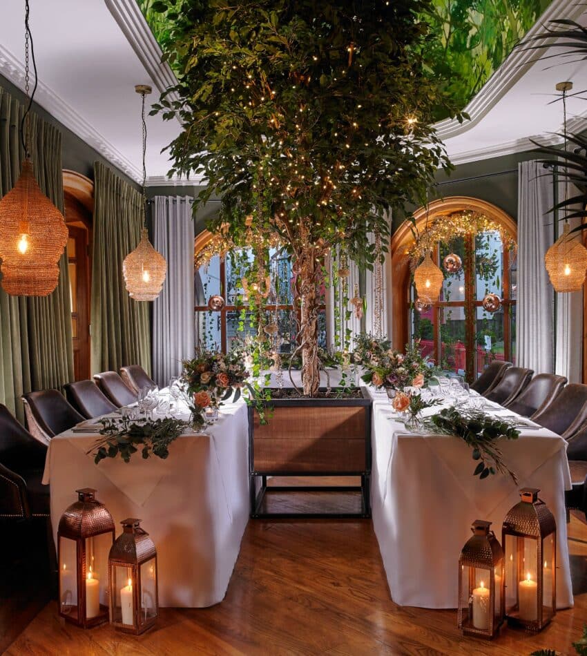 Elegant dining room with two tables arranged in an L-shape, decorated with greenery, candles, and lanterns, featuring a large indoor tree and ambient hanging lights.