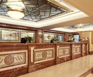 A hotel reception area with a marble front desk, decorative mosaic panels, flowers, and a glass-panelled ceiling.