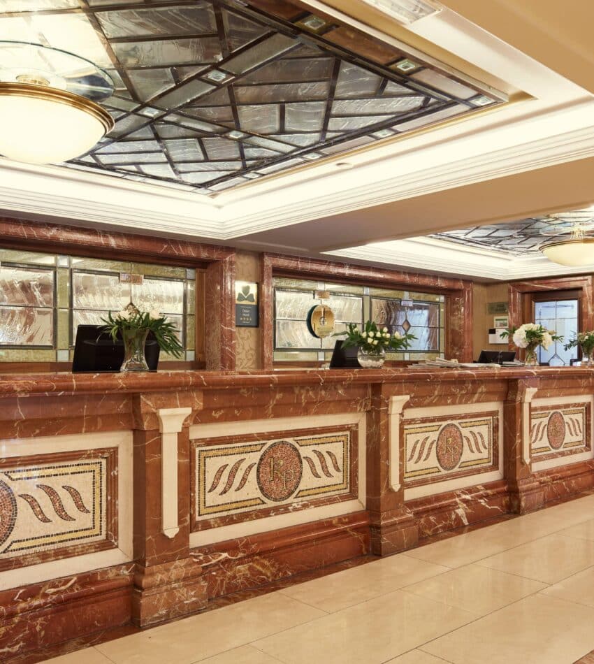 A hotel reception area with a marble front desk, decorative mosaic panels, flowers, and a glass-panelled ceiling.