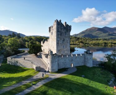 Stone castle with a central tower stands beside a lake, surrounded by green grass and trees, with mountains and blue sky in the background.